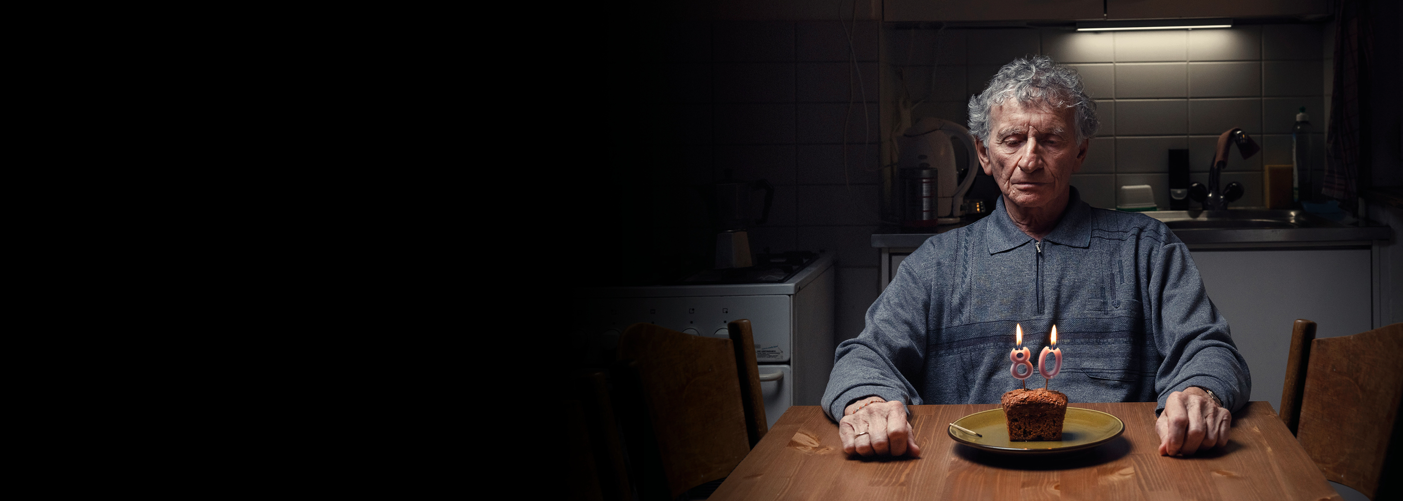 Ein Mann sitzt alleine vor einem kleinen Gebäck mit Geburtstagskerzen die die Zahl 80 zeigen. 